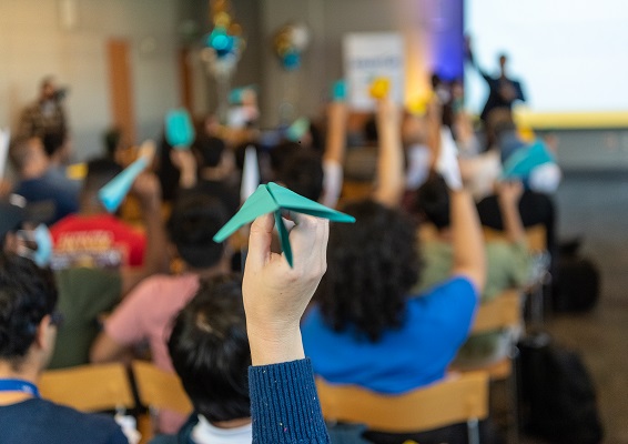 A roomful of people, seen from the back, hold up blue, white and gold paper planes, poised to throw toward an out-of-focus speaker at the front of the room A roomful of people, seen from the back, hold up blue, white and gold paper planes, poised to throw toward an out-of-focus speaker at the front of the room