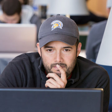 A student wearing a gray baseball cap ponders with his hand to his mouth while they stare at their laptop
